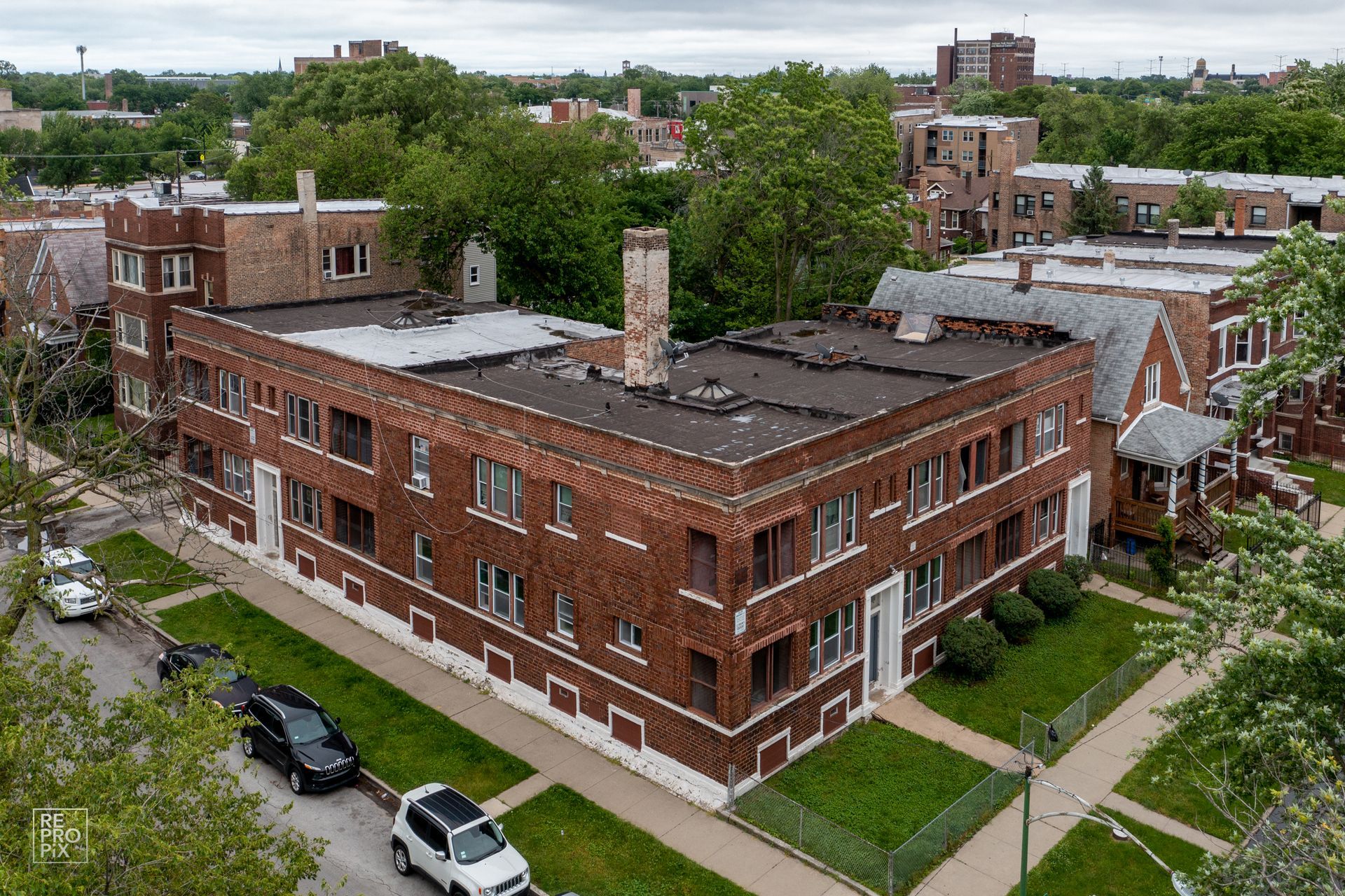 Brick apartment building with flat roof and chimney, trees, and parked cars on a city street.
