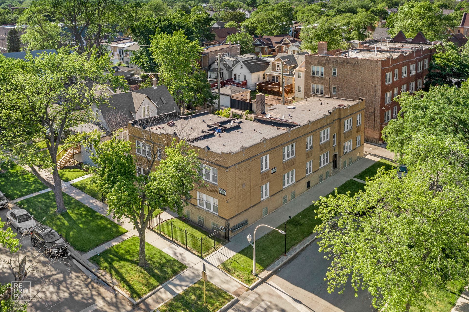 Aerial view of a two-story brick apartment building with surrounding trees and houses on a sunny day.