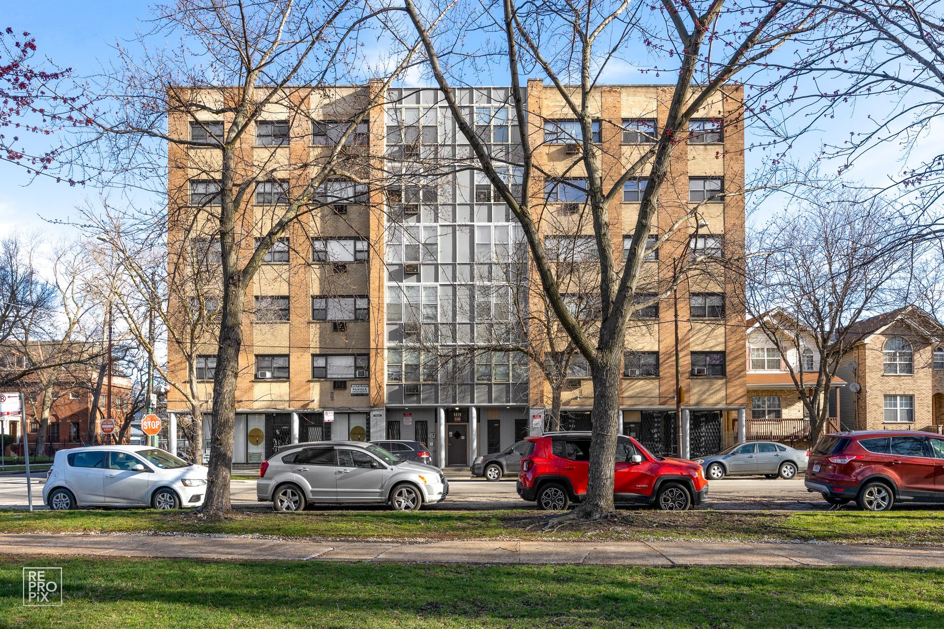 Apartment building facade with parked cars on a street; trees in front with a grassy area.