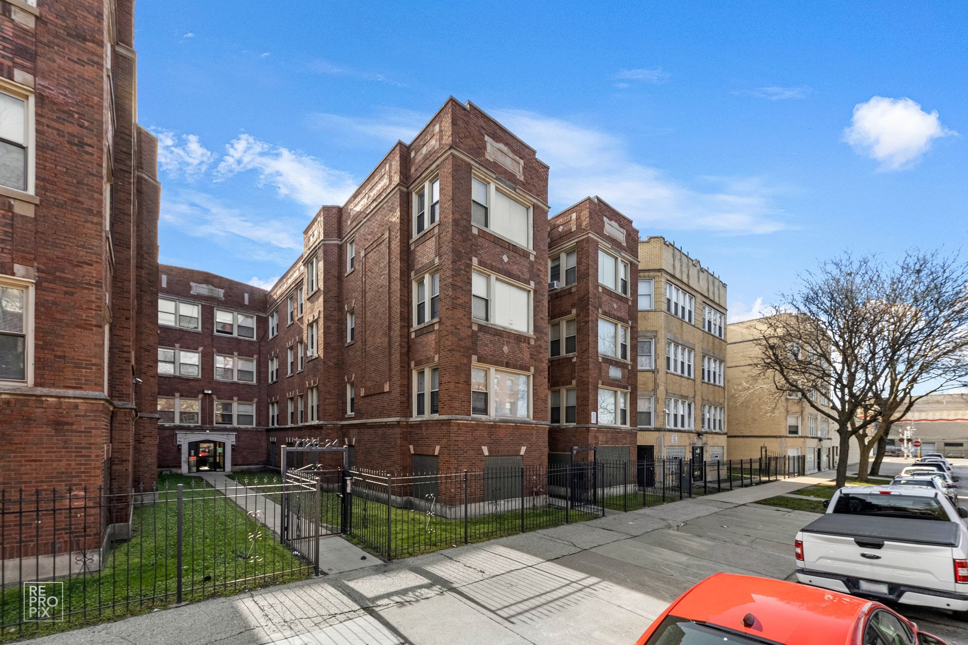 Brick apartment buildings with a small green lawn, and parked cars on the street under a blue sky.