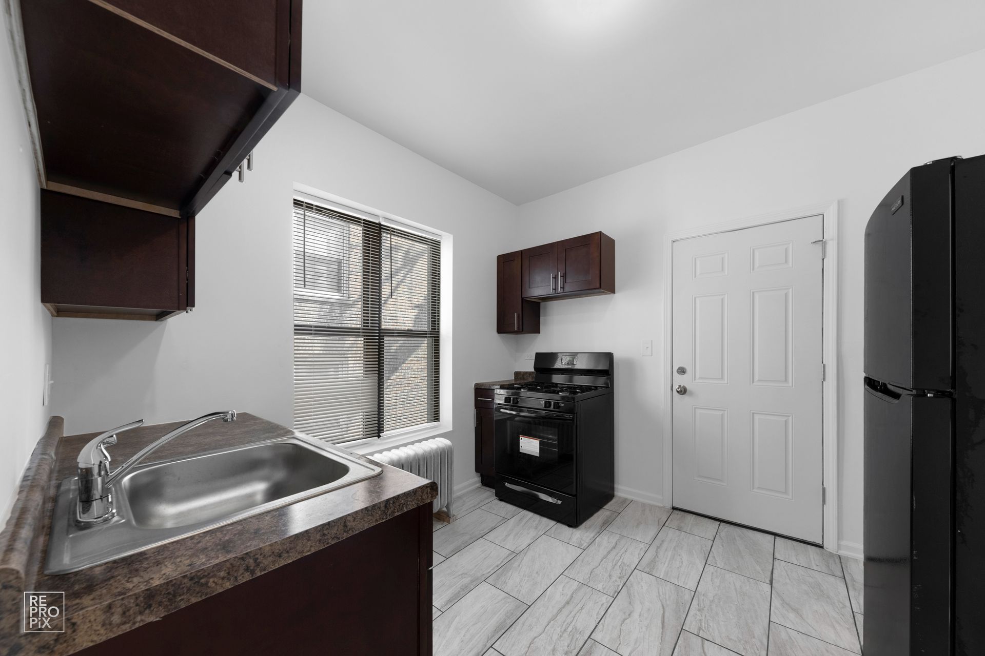 Small kitchen with dark brown cabinets, black appliances, white door, and grey floor tiles.