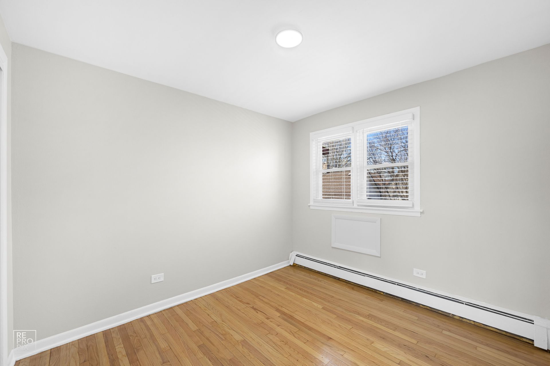 Empty room with light gray walls, wood floor, window, and radiator.