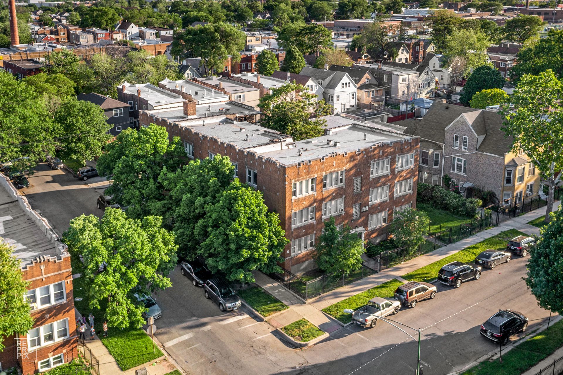 Aerial view of brick apartment building with surrounding trees and parked cars.
