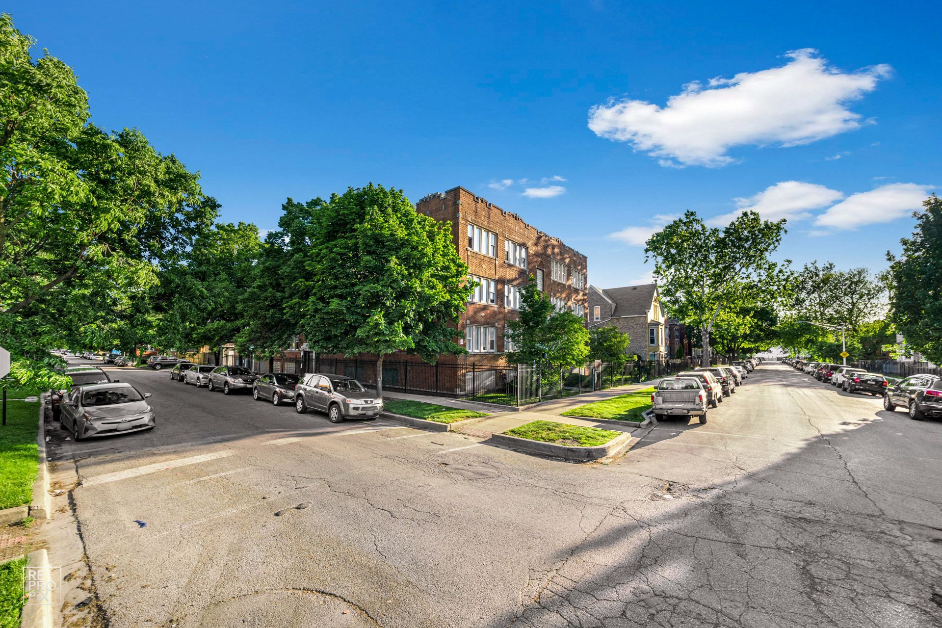 Corner of a city street with parked cars, apartment building, trees, and blue sky.
