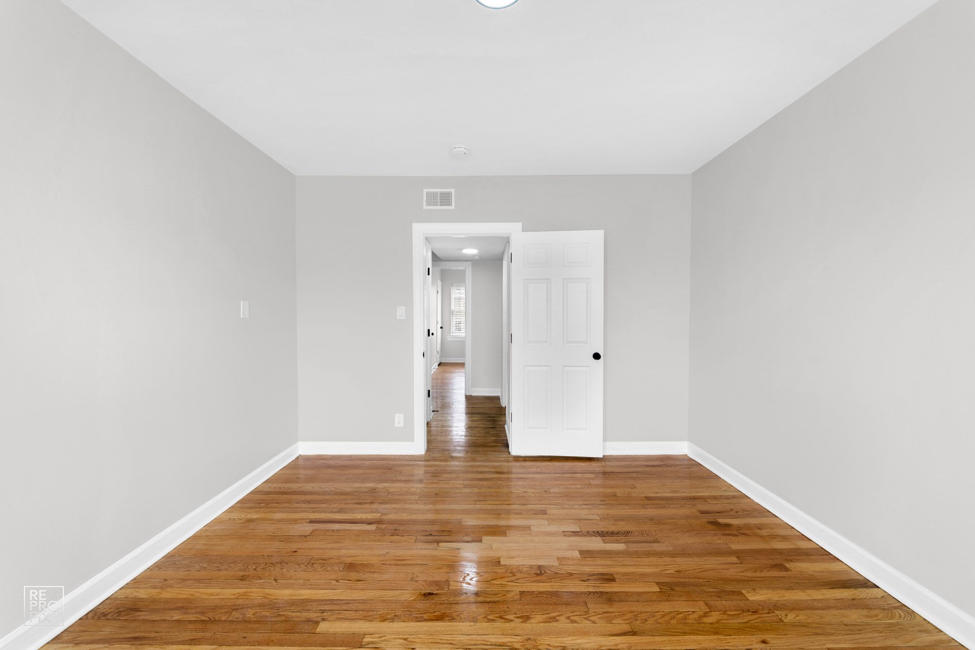 Empty room with hardwood floors, light gray walls, and white trim. A doorway leads to a hallway.