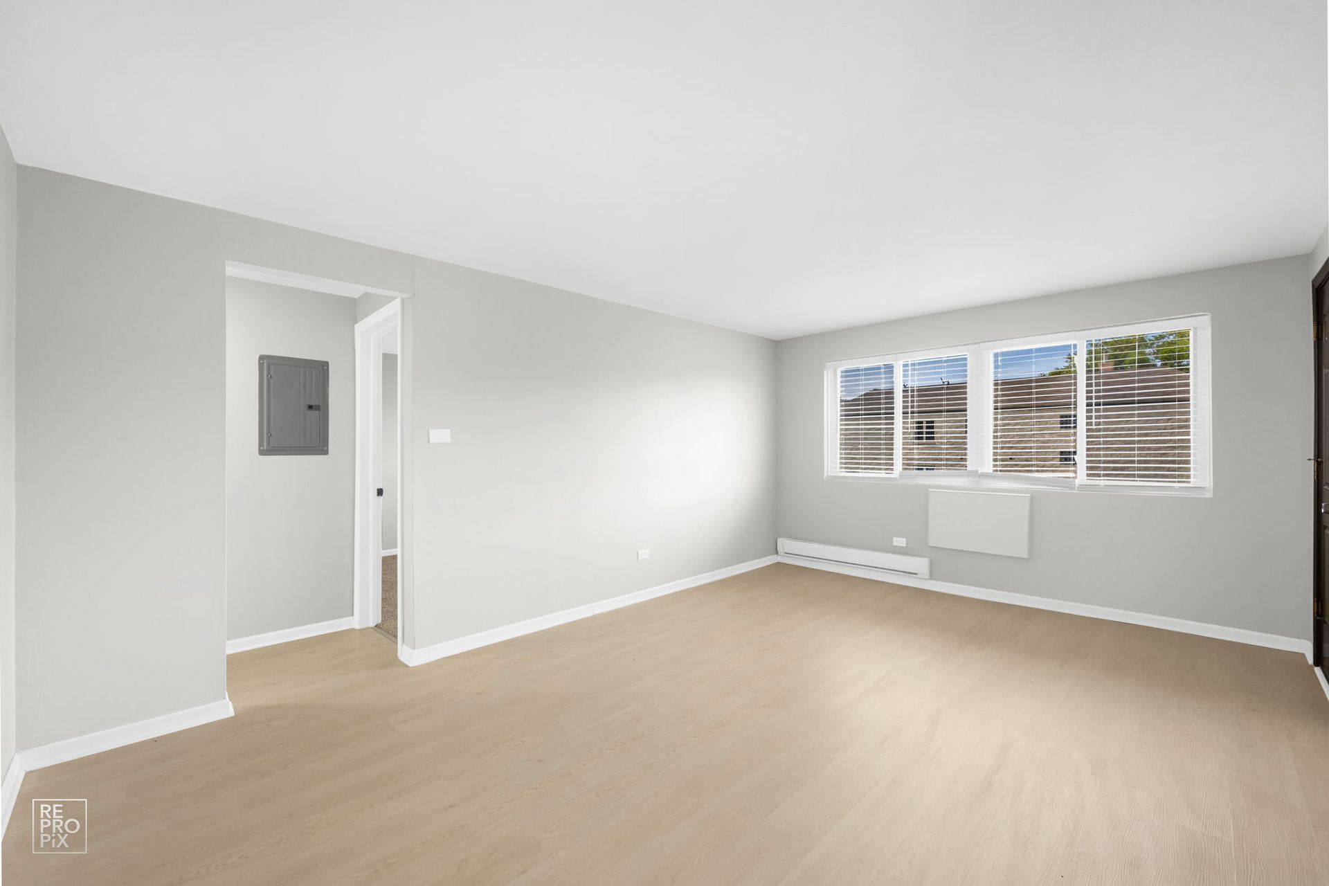 Empty, light-filled living room with beige carpet, white walls, and a window. An open doorway leads to another room.