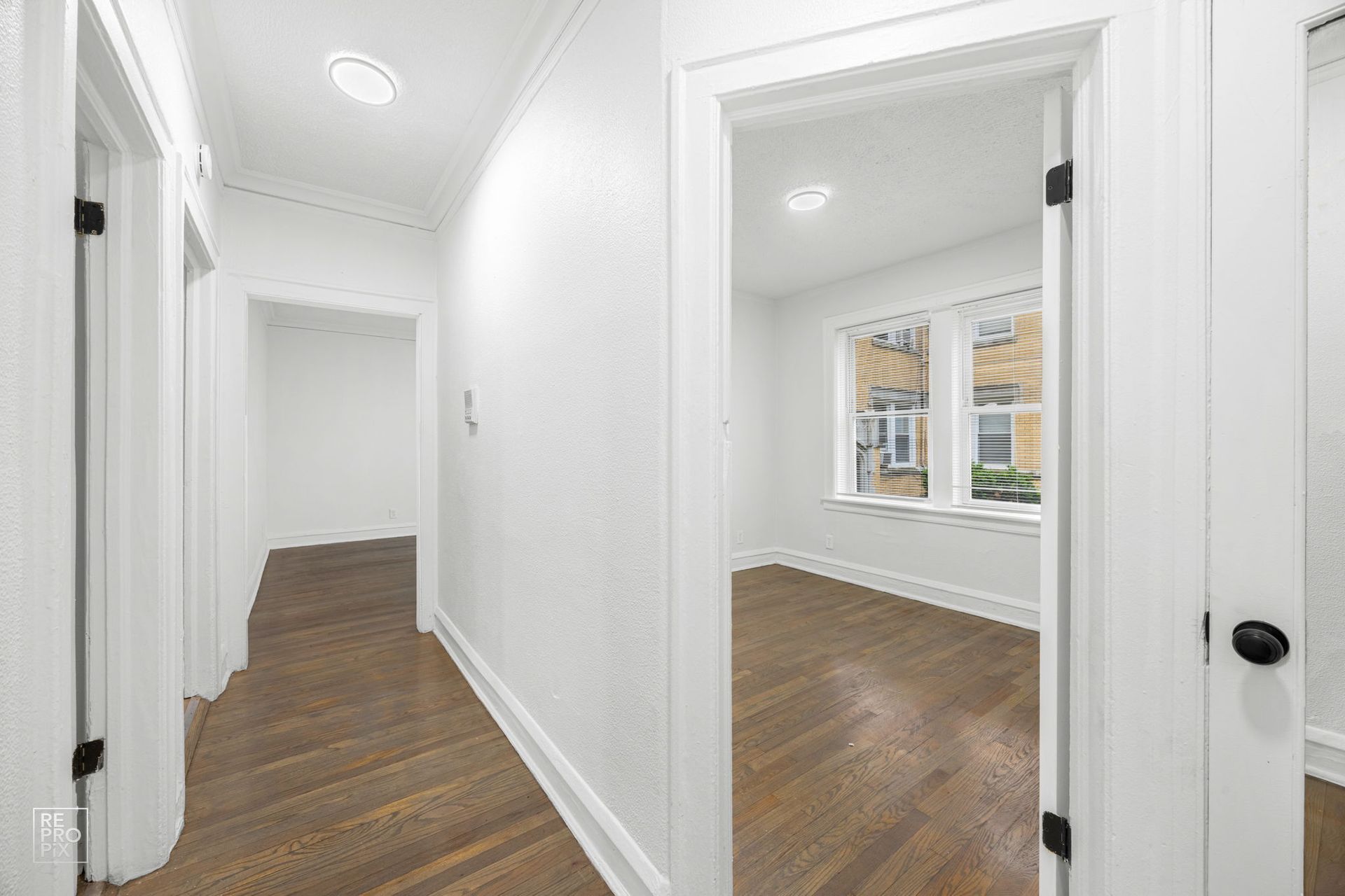 Bright white hallway with wooden floors; doorway leads to room with window.