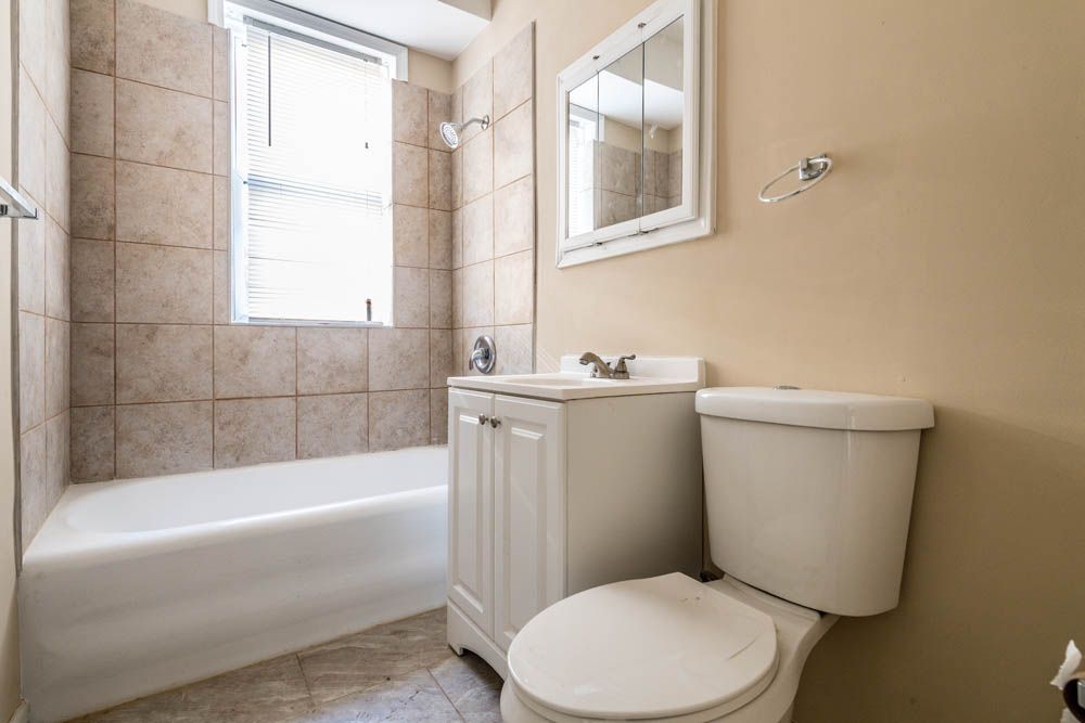 Small bathroom with a white tub, toilet, and vanity. Beige tile and walls, a window and a mirror.