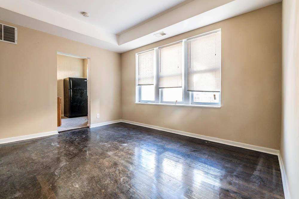 Empty room with dark floor, beige walls, windows with blinds, and doorway to a kitchen with a black refrigerator.