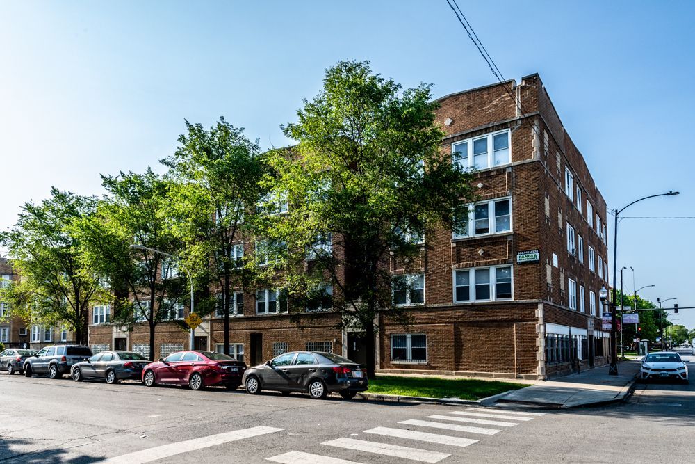 Apartment building on a city street, brick exterior, parked cars, trees, blue sky.