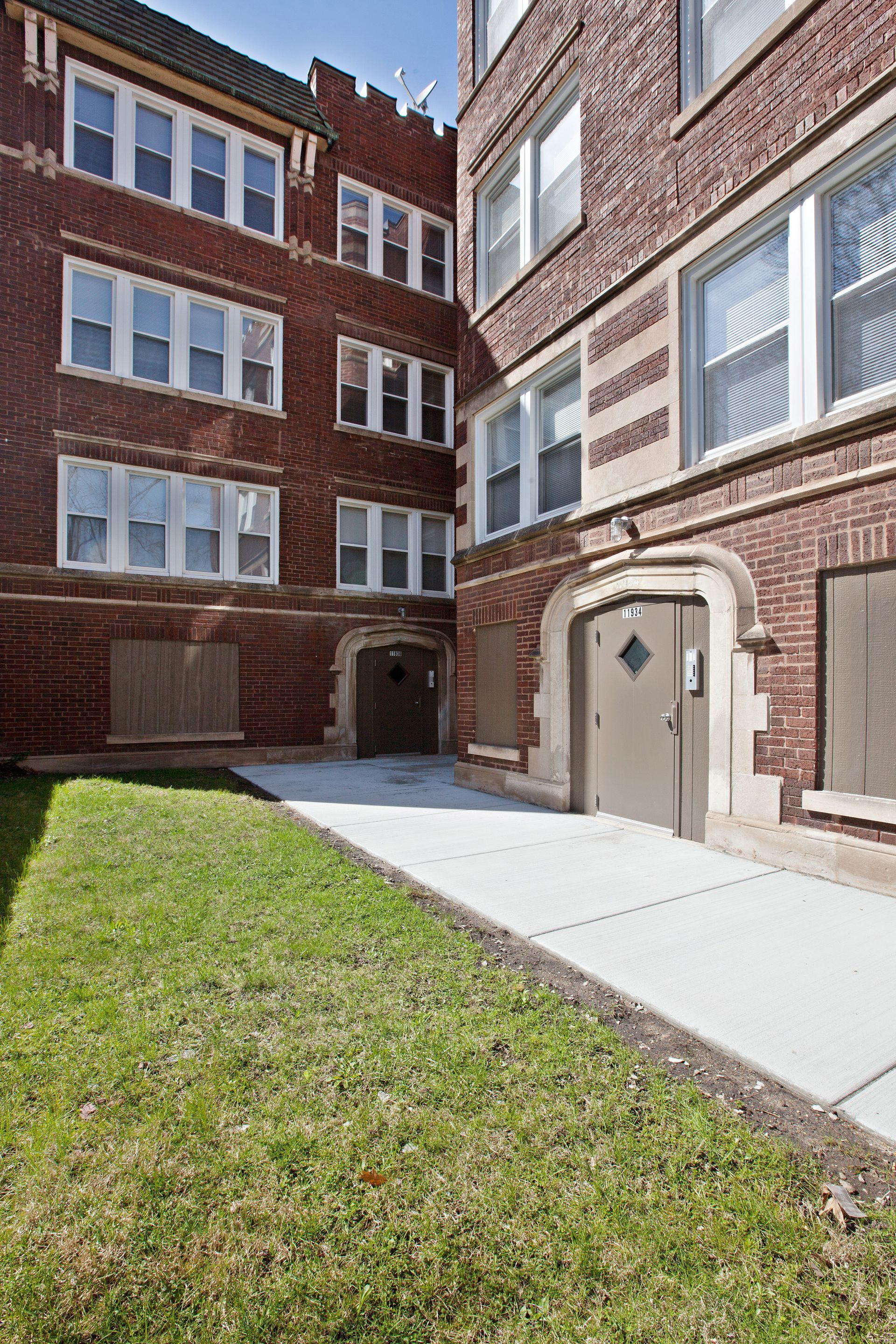 Apartment building exterior, brick facade, arched entryway, sidewalk, grassy area, sunny day.