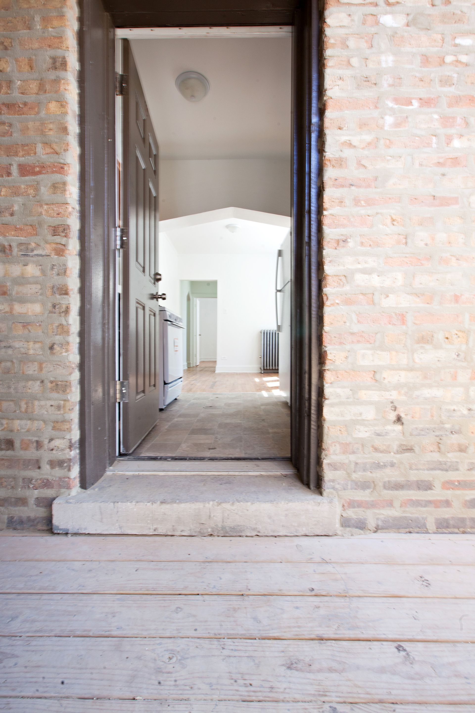 Open brown door into a room with exposed brick walls, light wood floor, and white walls.
