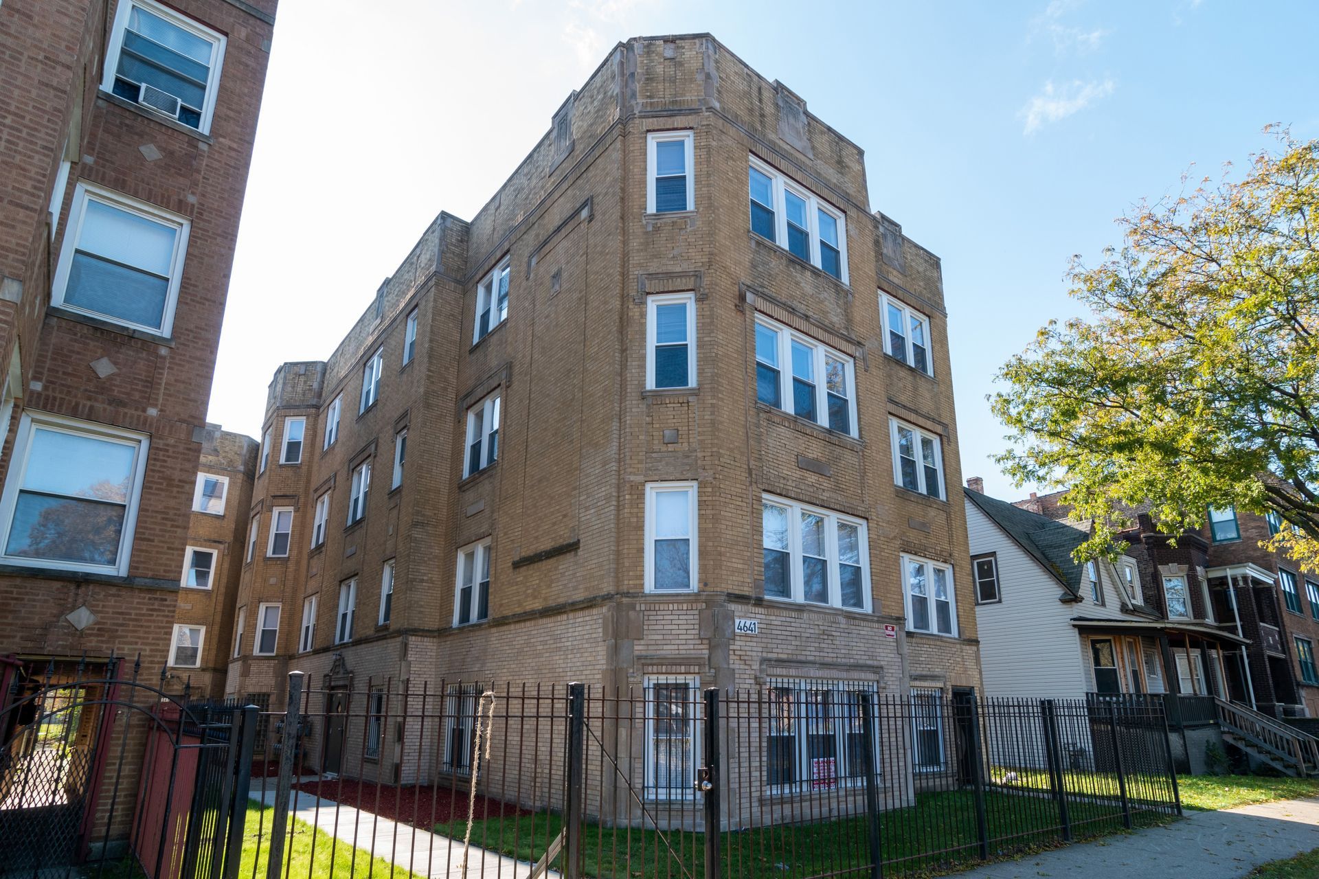Three-story brick apartment building behind a black metal fence on a sunny day.