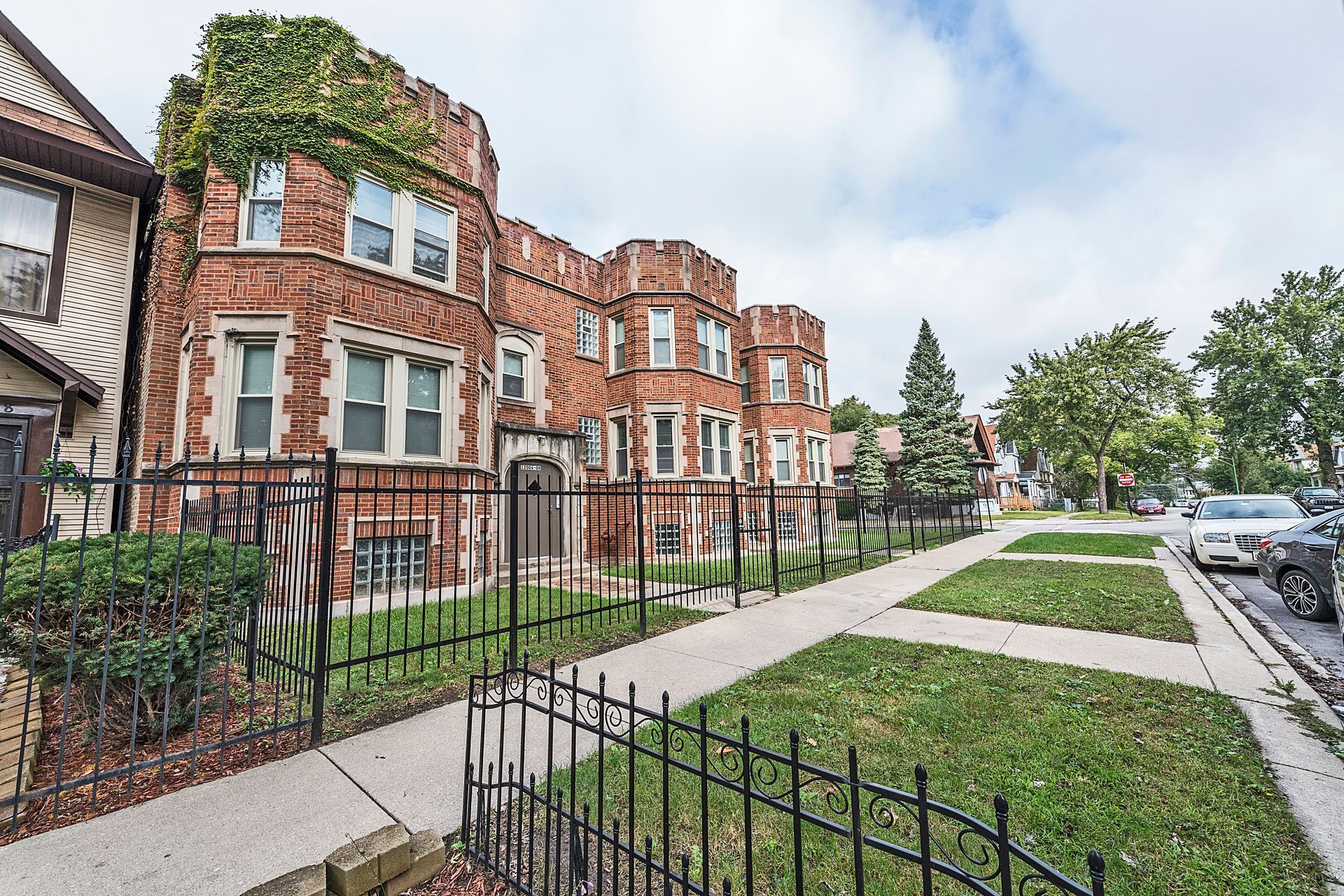 Brick apartment building with a wrought-iron fence and sidewalk. Overgrown with green ivy.