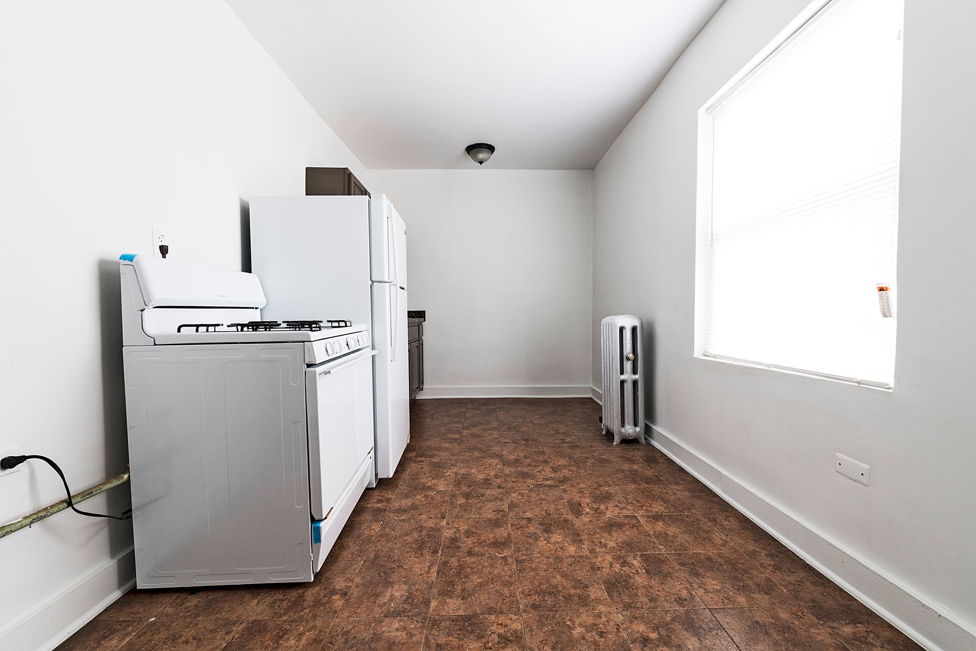 Small kitchen with white appliances, brown flooring, and a window with blinds.