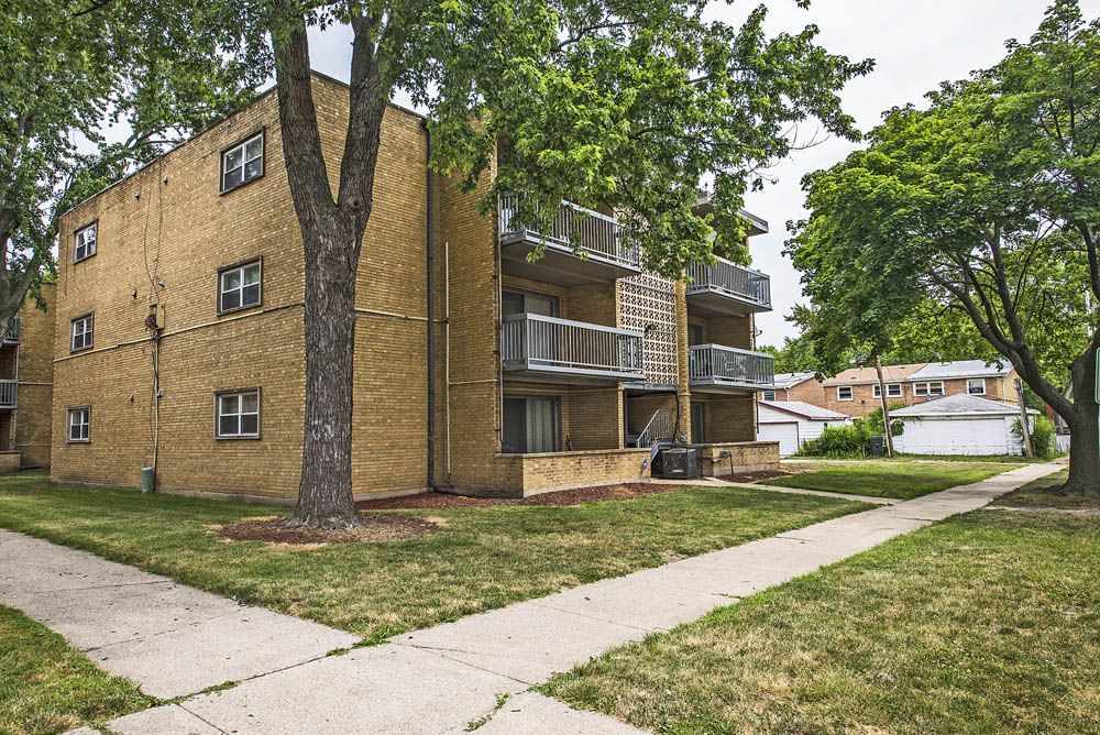 Three-story brick apartment building with balconies, trees, and sidewalks on a sunny day.