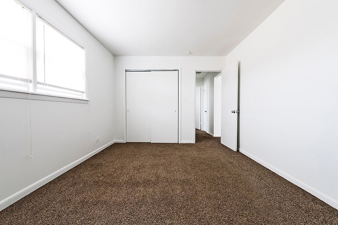 Empty room with white walls, brown carpet, window with blinds, and closet.
