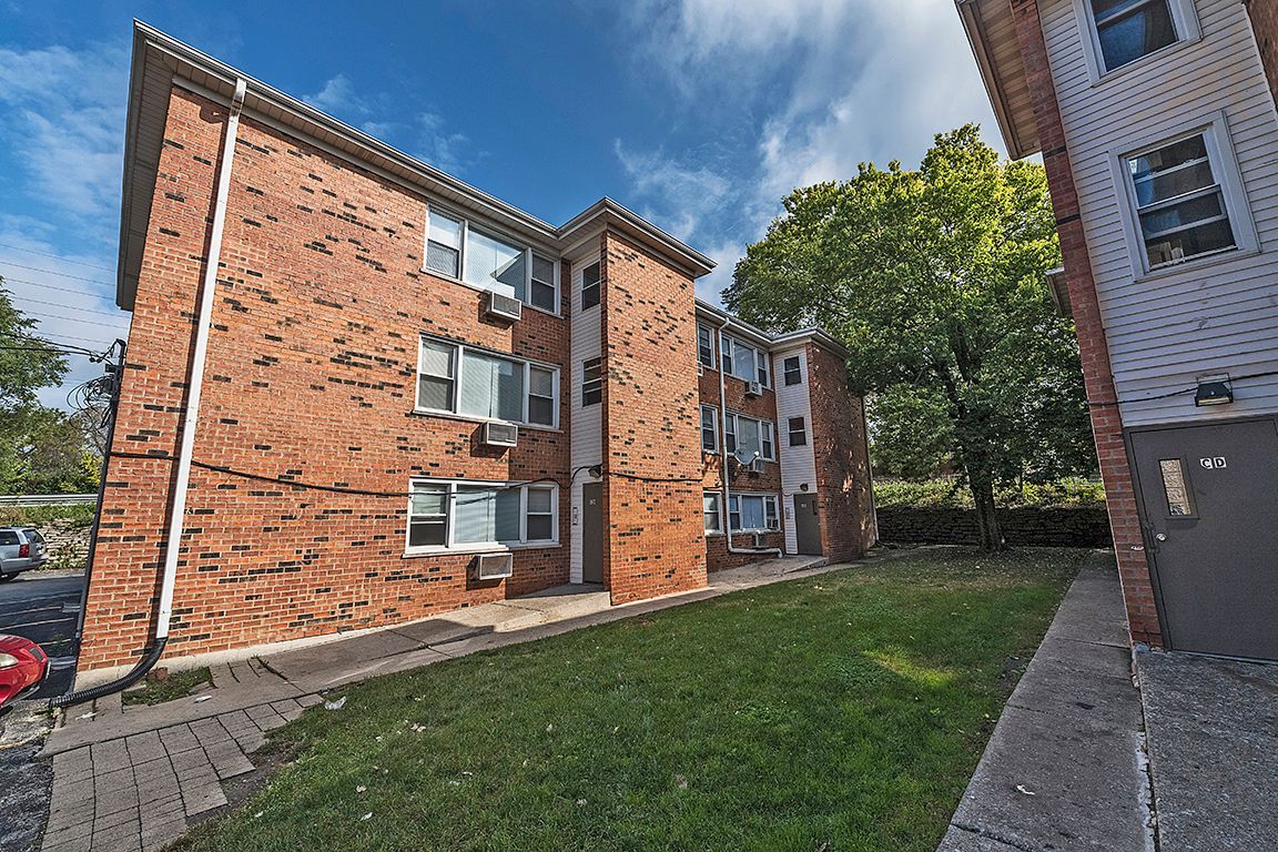 Brick apartment building with green lawn under a blue sky, some parked cars are visible.