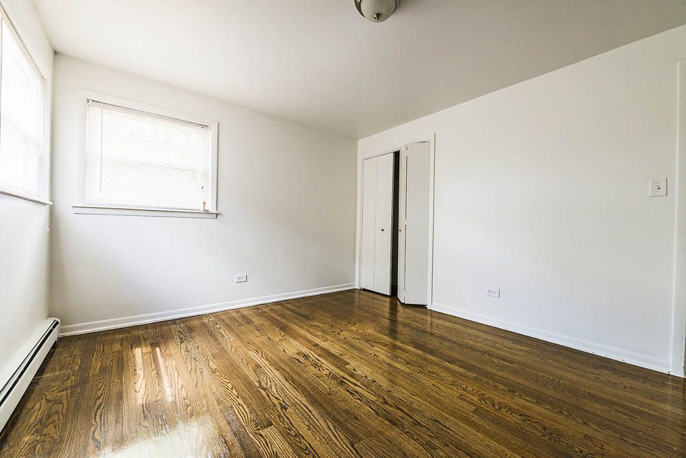 Empty bedroom with hardwood floors, white walls, closet, and a window with blinds.