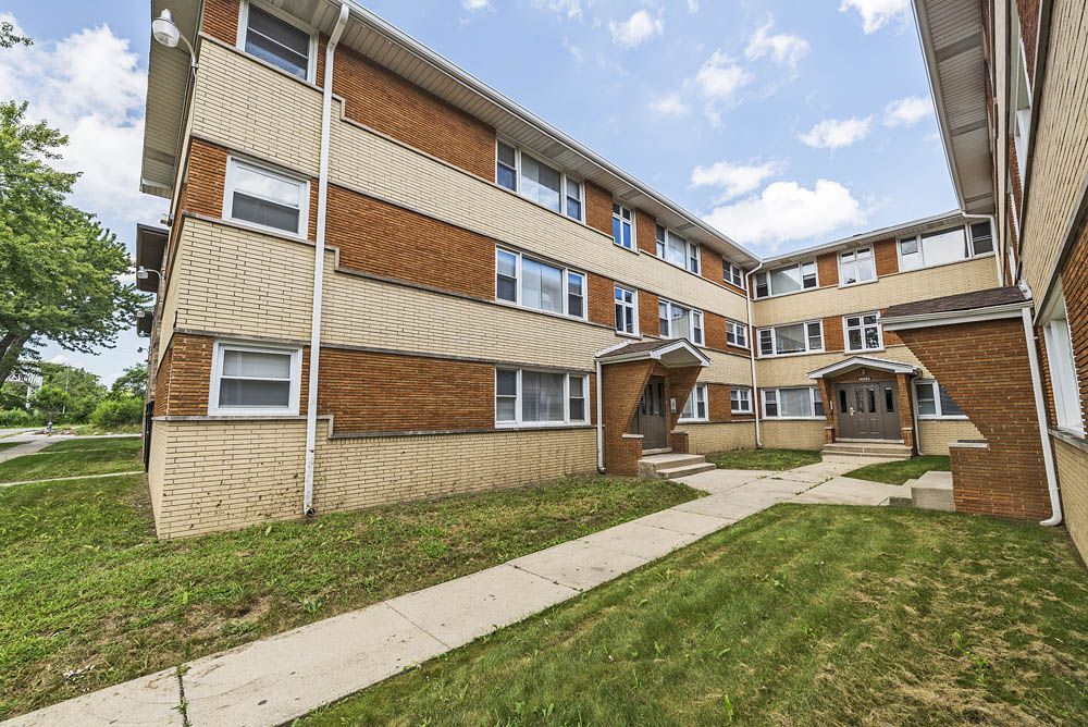 Three-story brick apartment building with beige siding, windows, and green lawn.