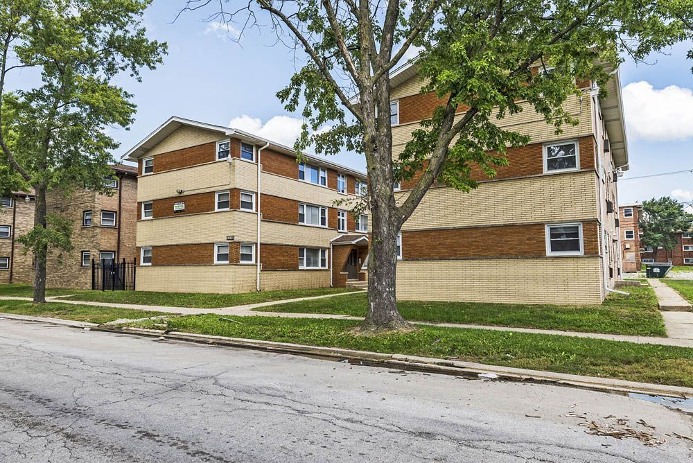 Apartment building with brick and tan siding, surrounded by grass and trees on a sunny street.