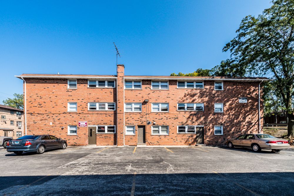 Red brick apartment building with parking, cars parked in front, and a blue sky.