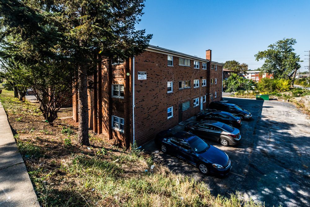 Brick apartment building with parked cars, surrounded by trees and overgrown vegetation.