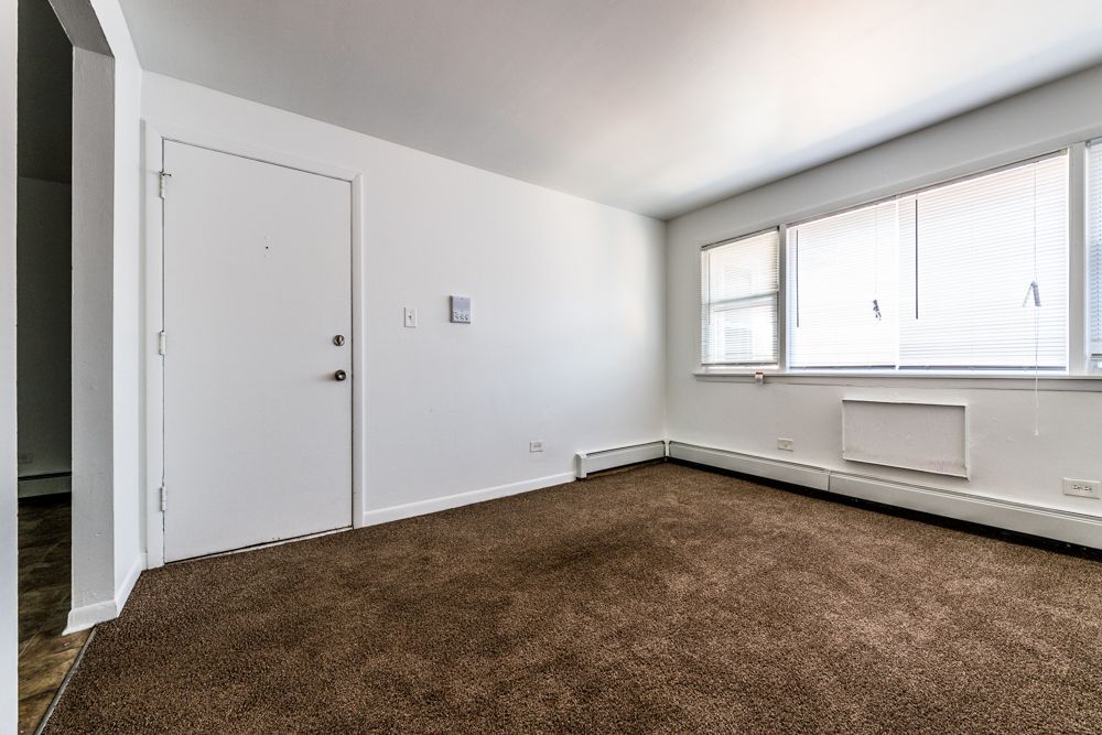 Empty living room with brown carpet, white walls, and a window. A door is on the left.