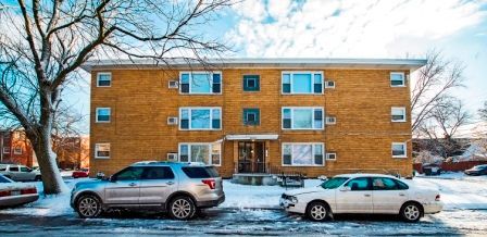 A two-story brick apartment building on a snowy street with a silver SUV and white car parked in front.