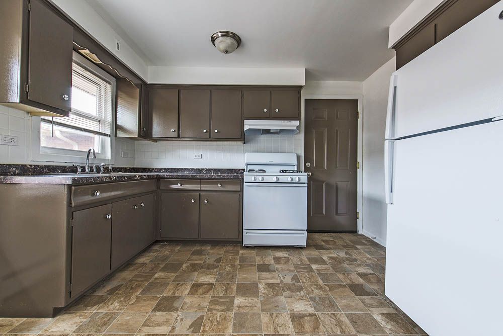 Brown kitchen cabinets, white appliances, and brown patterned flooring in an empty room.