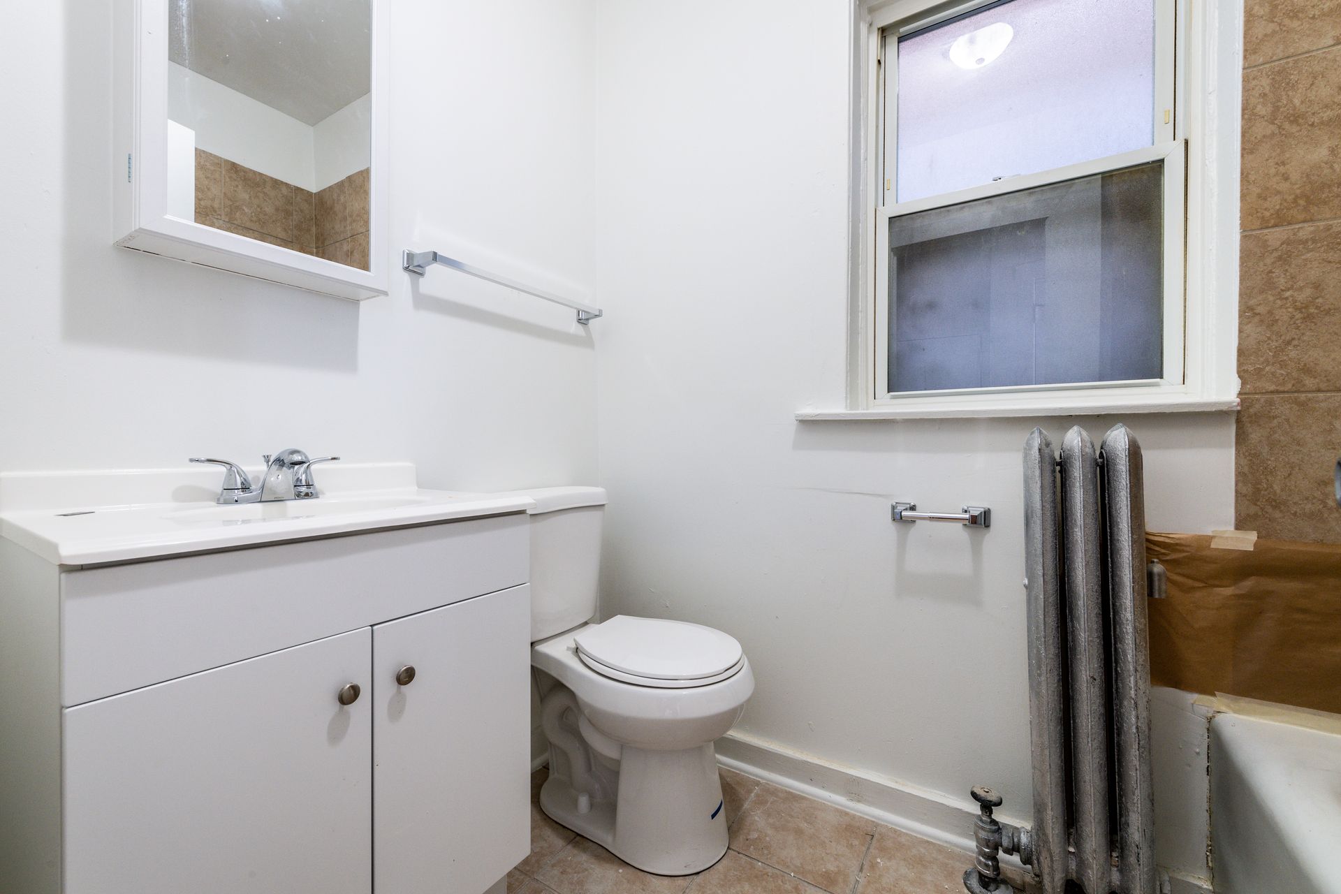 Small white bathroom with toilet, sink, mirror, window, and radiator.