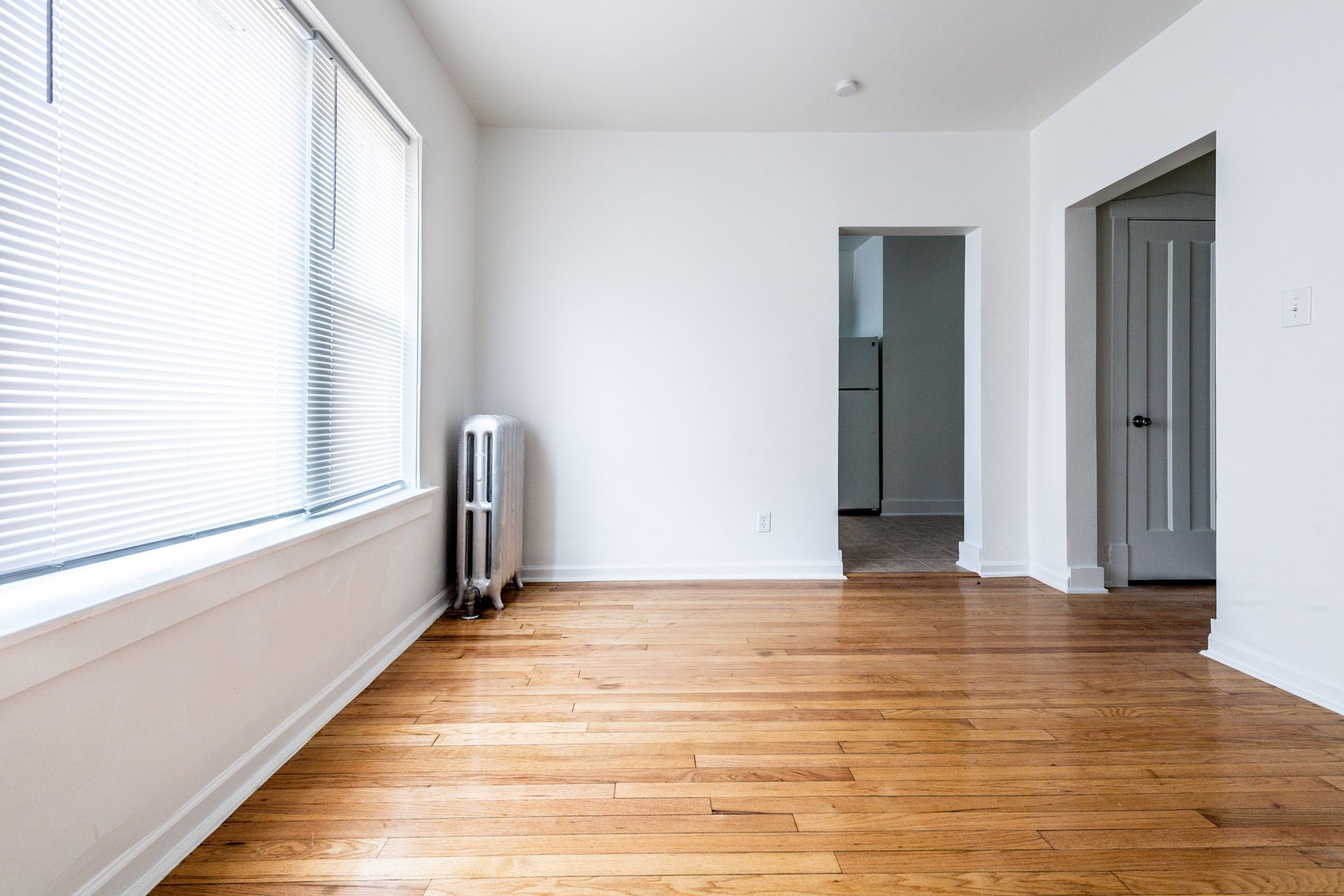 Empty room with wood floors, large window with blinds, radiator, and doorways.
