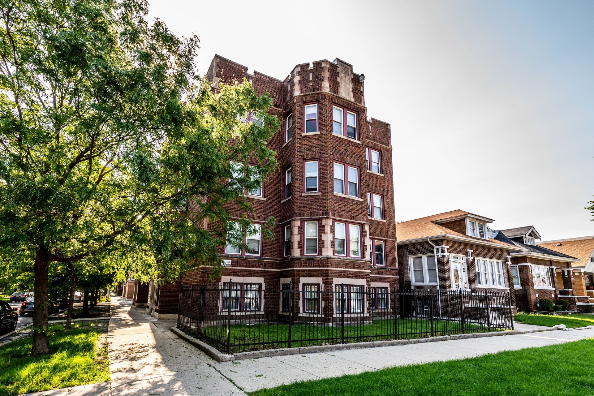 Brick apartment building with decorative turret, green lawn, sidewalk, and nearby houses.