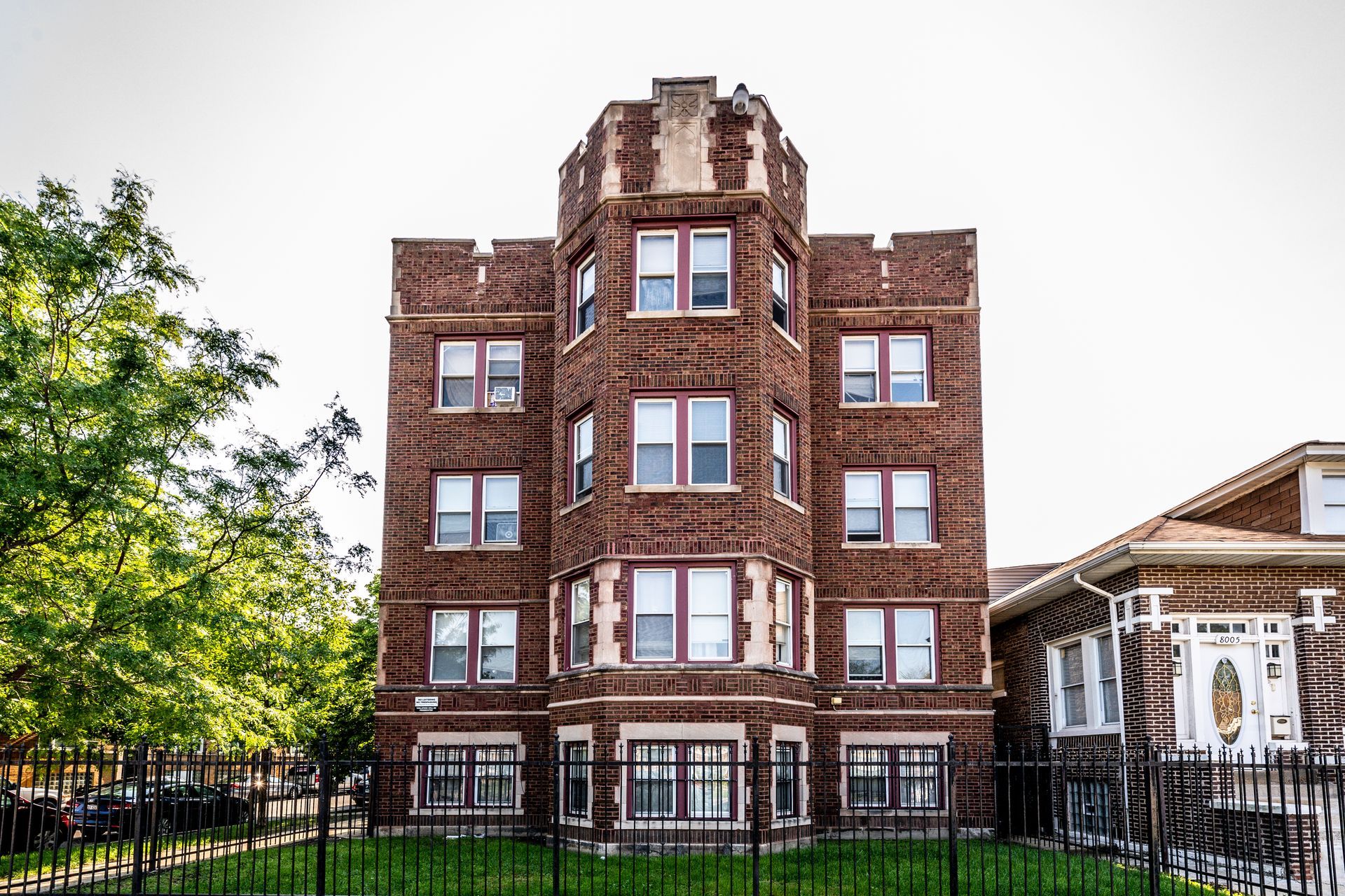 Brick apartment building with turret-like structure and multiple windows, with green trees and a fence in front.