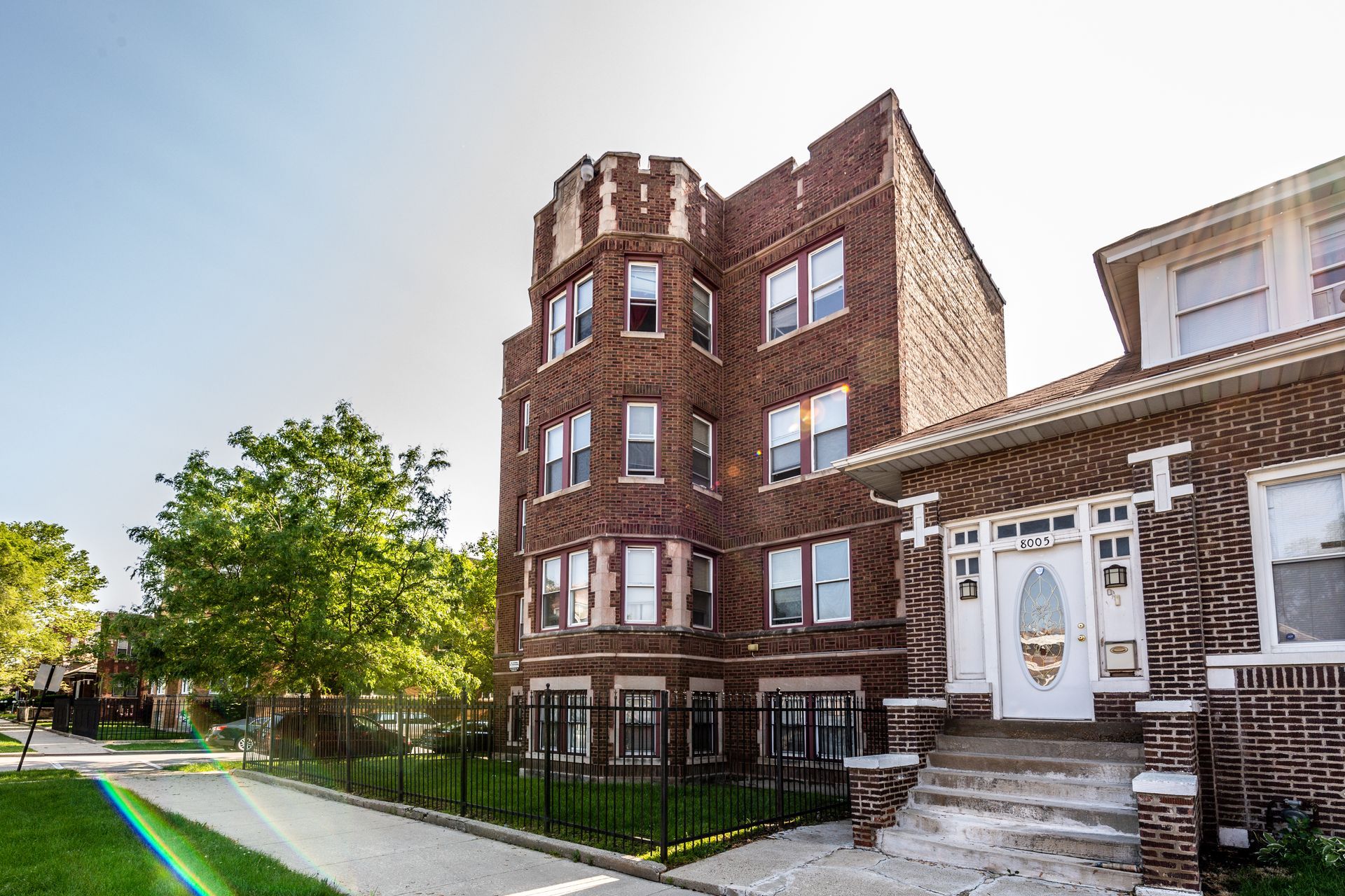 Brick apartment building with turret and arched doorway, blue sky, and green lawn.