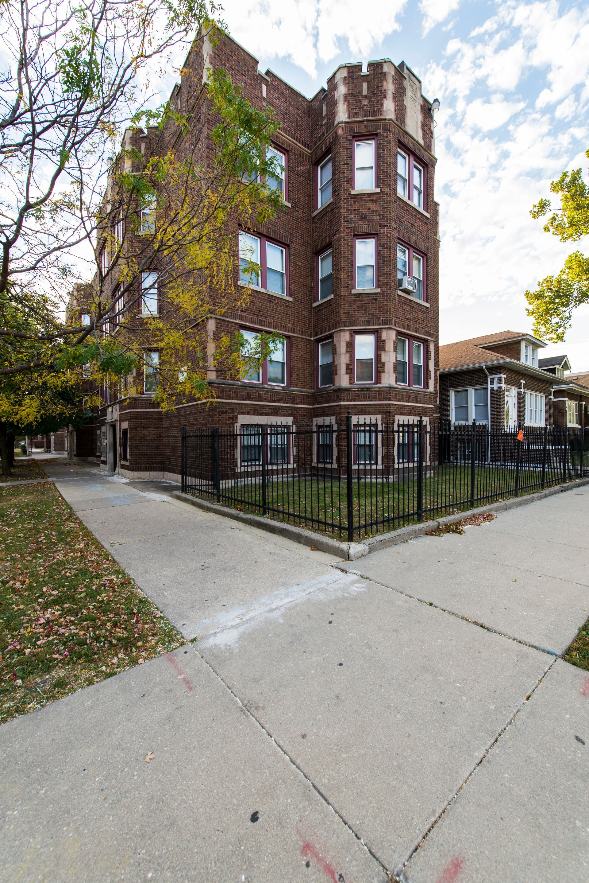 Brick apartment building on a corner lot, surrounded by a black fence and sidewalk, with a tree in fall colors.