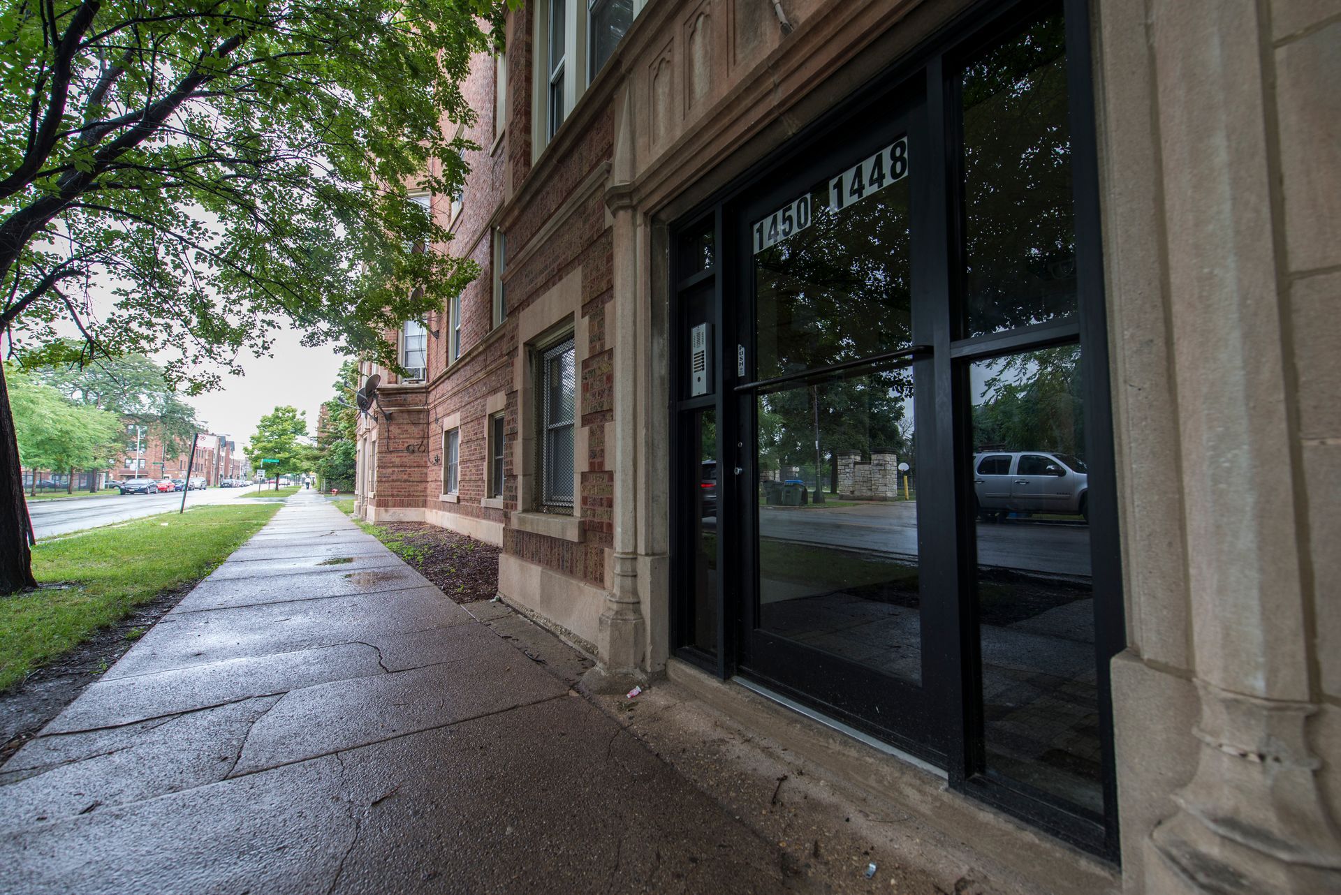 Exterior of a building with a black-framed storefront and a wet sidewalk. Trees line the street on a cloudy day.