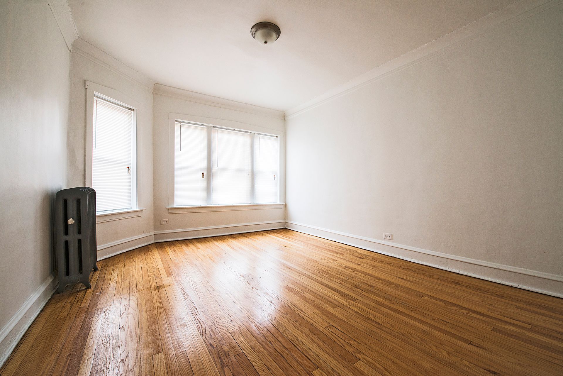 Empty room with hardwood floors, white walls, and a radiator. Windows with blinds.