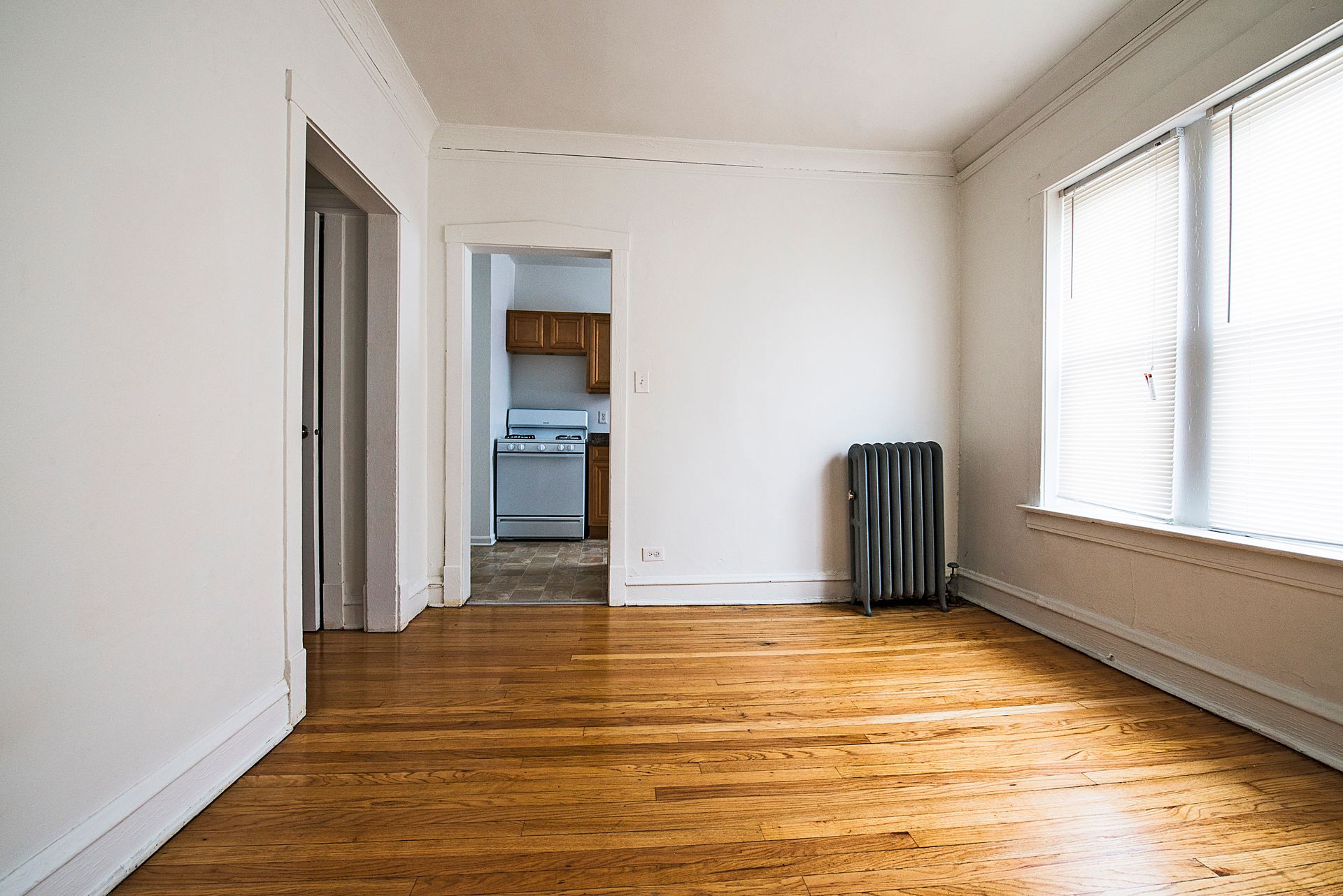 Empty room with hardwood floors, white walls, and a window with blinds. Doorways lead to kitchen and hall.