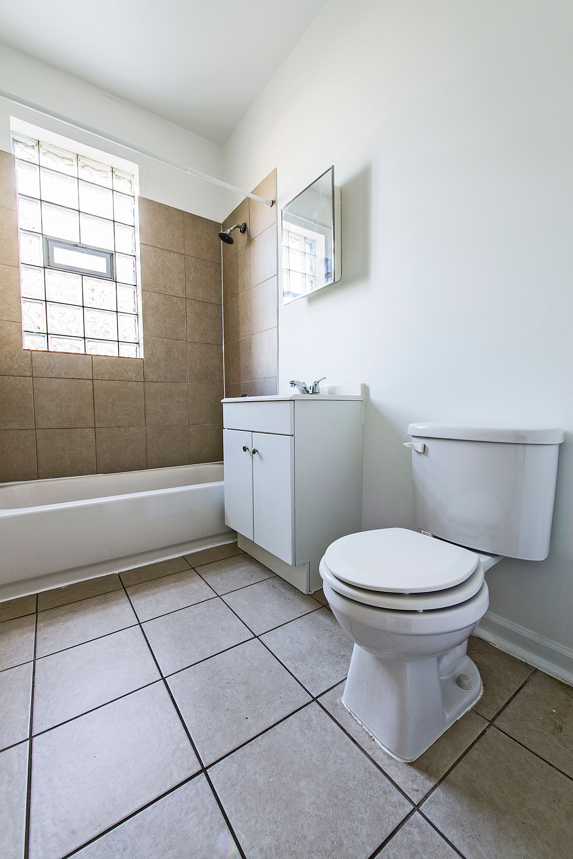 Bathroom with white toilet, sink, and tub. Beige tiled walls and floor. Window on the left.