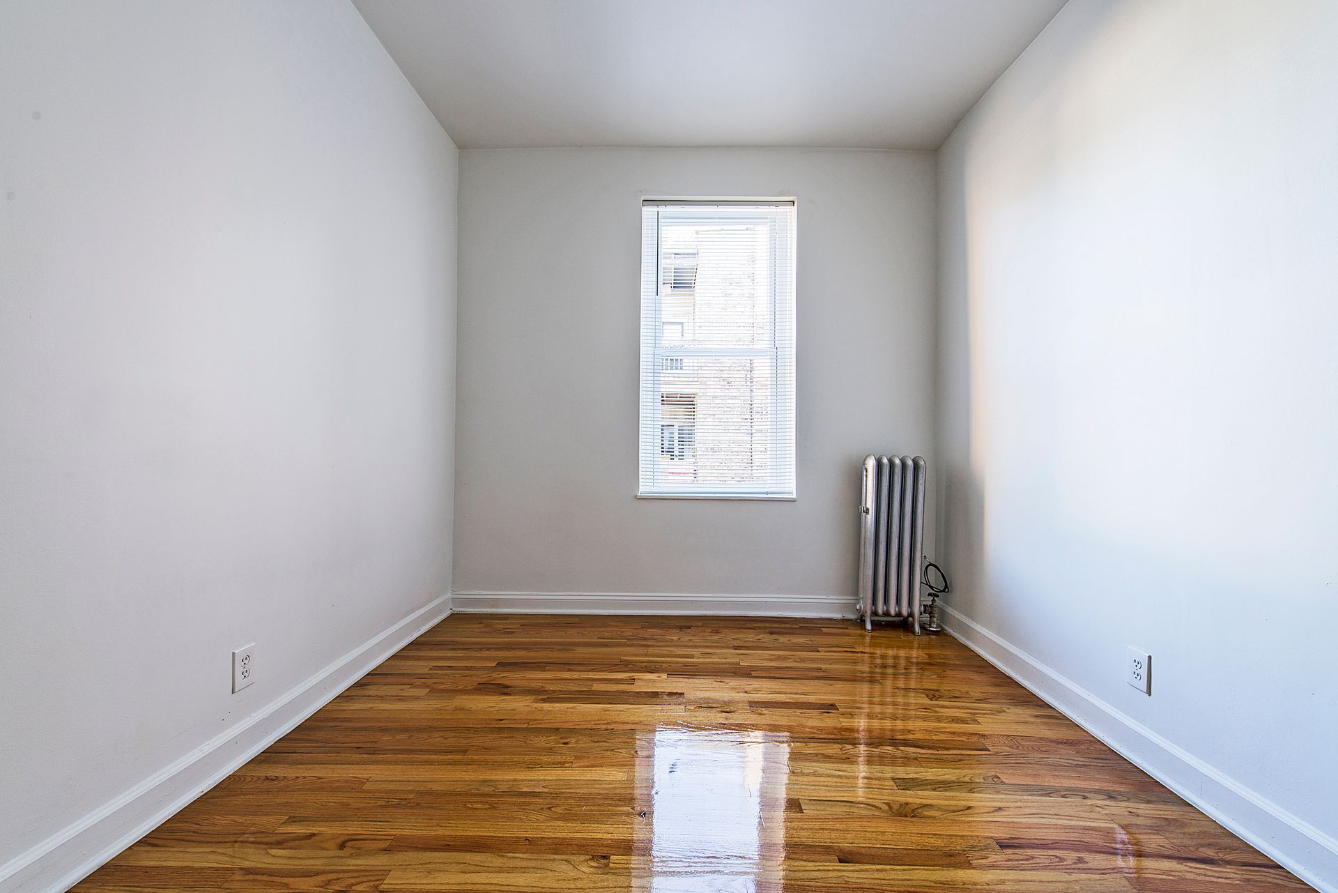 Empty room with hardwood floors, a window with blinds, and a radiator. White walls.