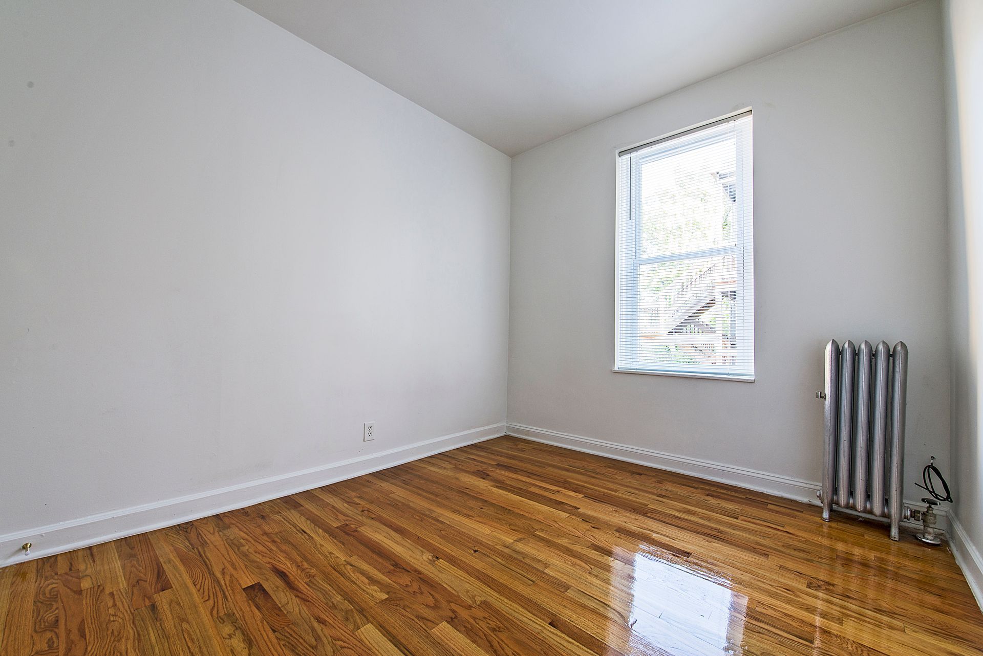 Empty room with wood floor, white walls, radiator, and window with blinds.