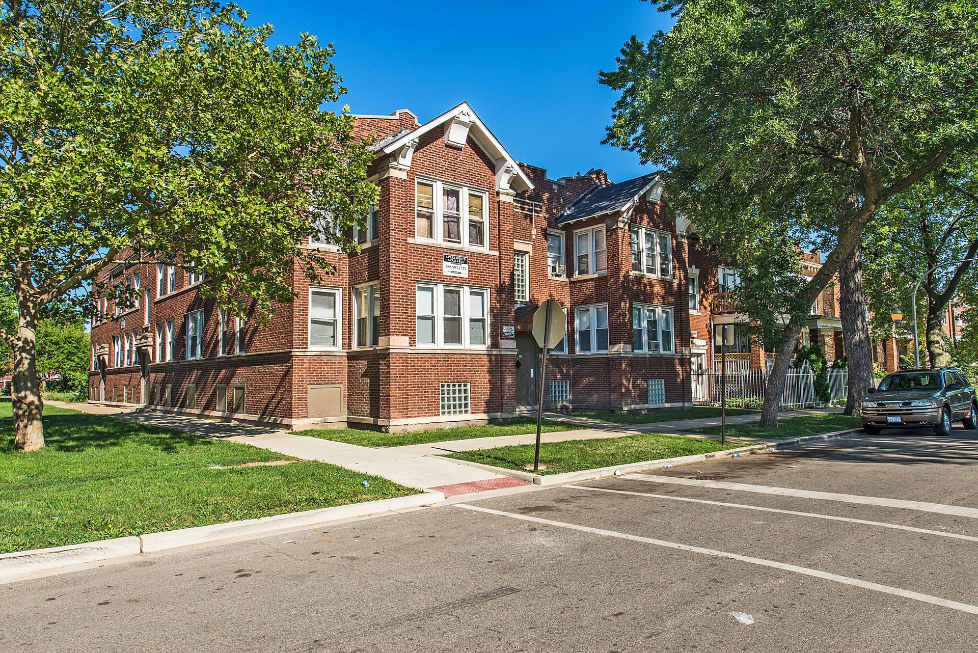 Brick multi-unit building on a corner lot with trees and a parked car. Sunny day.