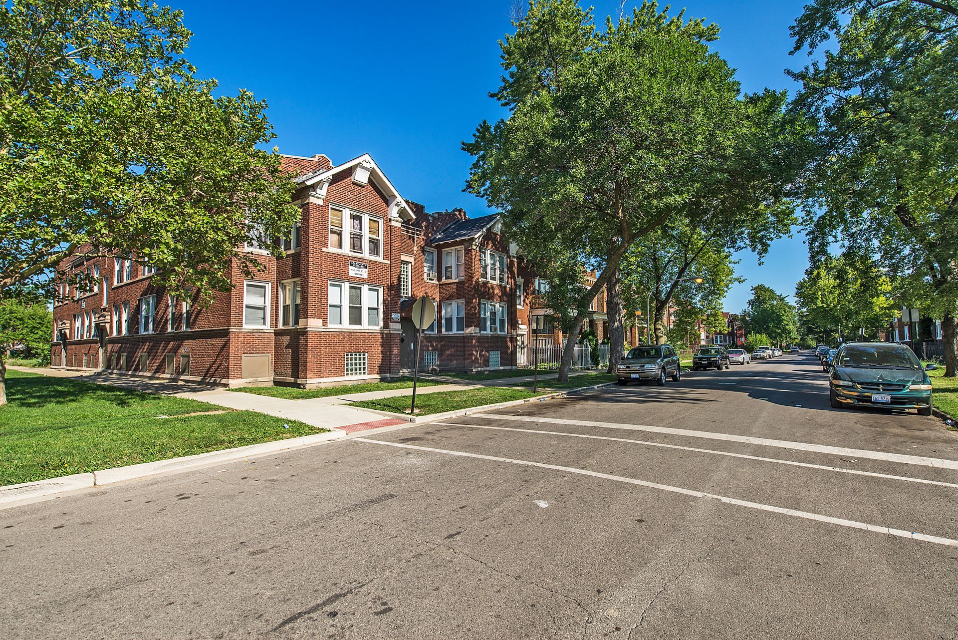 Brick apartment building on a tree-lined street under a bright blue sky.