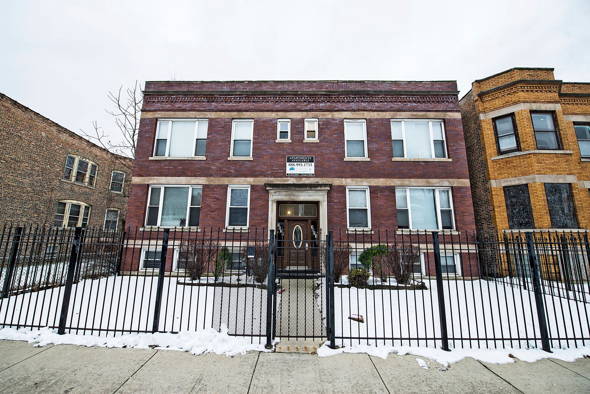 Red brick apartment building with black wrought-iron fence; snow on the ground; cloudy sky.