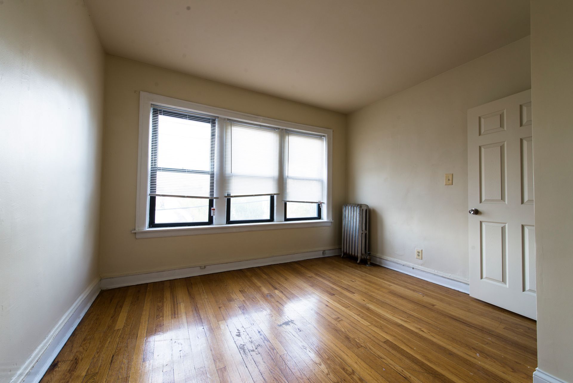 Empty room with hardwood floors, a window with blinds, and a closed door.