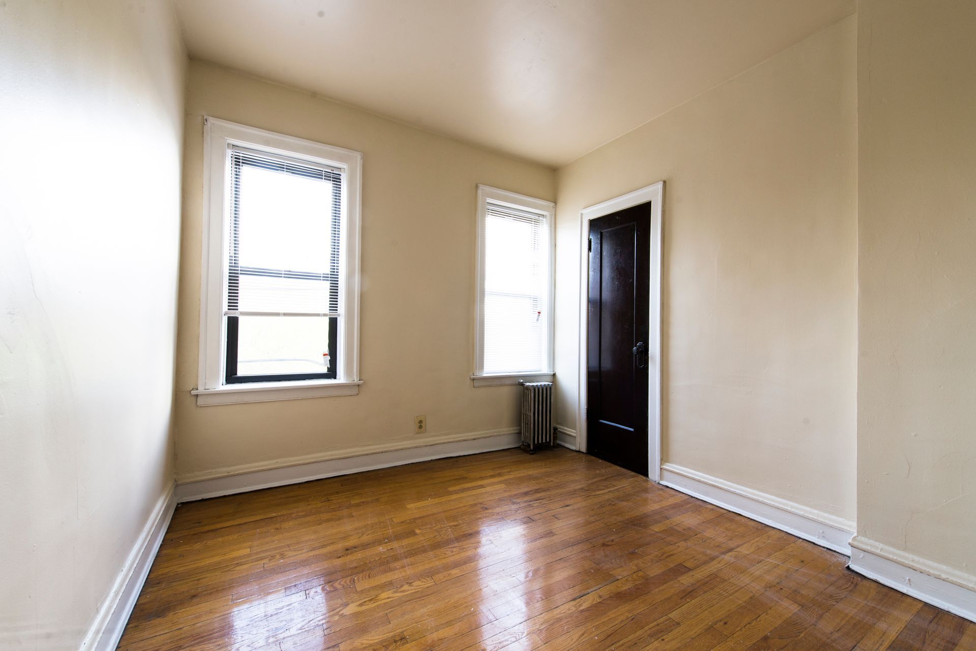 Empty room with hardwood floors, two windows, and a dark door. Pale walls, white trim.