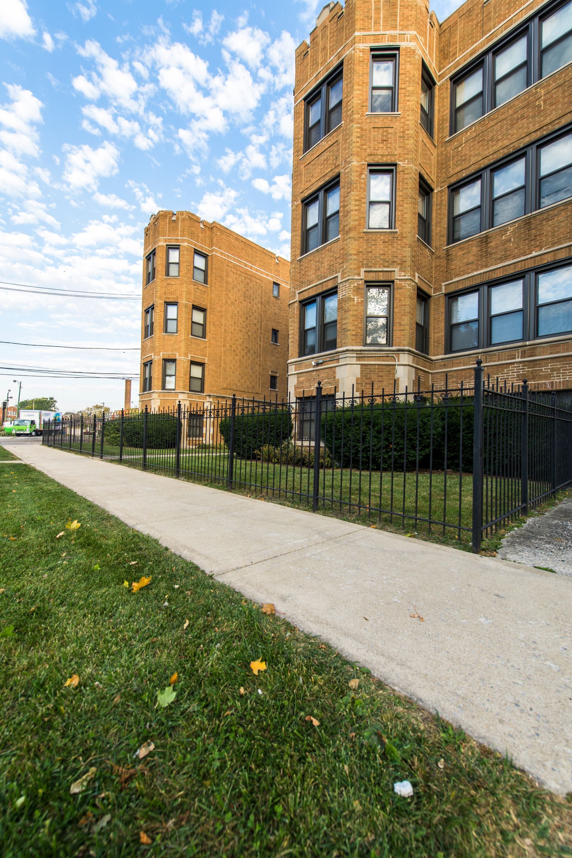 Brown brick apartment buildings, black fence, sidewalk, green grass, blue sky.