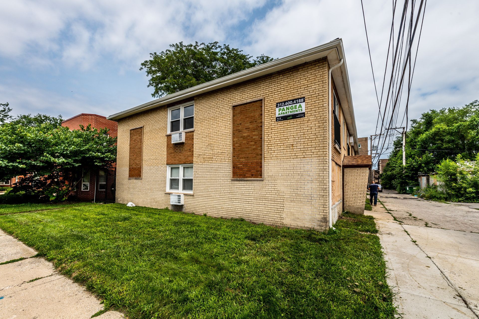 Brick building with boarded-up windows and overgrown lawn on a sunny day. Power lines visible.