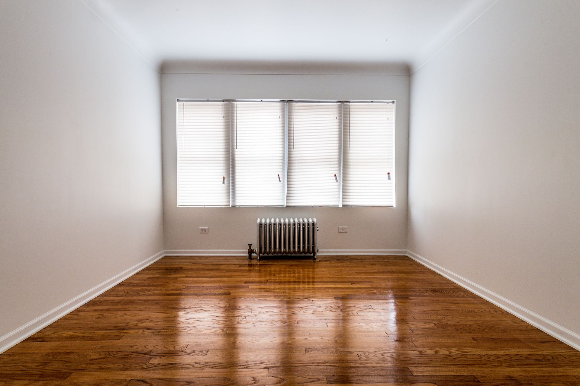 Empty room with hardwood floors, a window with blinds, and a radiator.